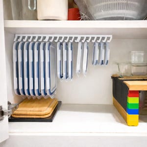 May include: A white kitchen cabinet interior with a hanging storage system for food containers. Blue and white rectangular containers hang from hooks. A wooden chopping board sits on the bottom shelf. Other kitchen items are visible.