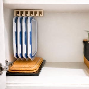 May include: A beige plastic storage rack mounted under a shelf, holding four rectangular food container lids with white frames and blue silicone seals. Below the lids, two stacked rectangular bamboo cutting boards rest on a black base.