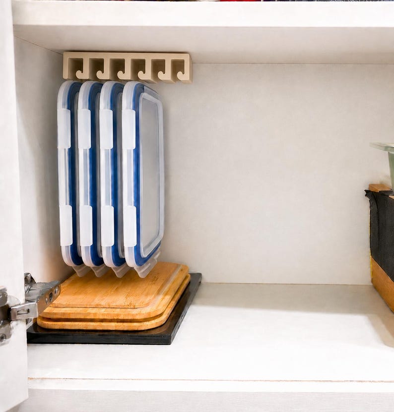 May include: A beige plastic storage rack mounted under a shelf, holding four rectangular food container lids with white frames and blue silicone seals. Below the lids, two stacked rectangular bamboo cutting boards rest on a black base.