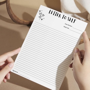 May include: A lined notepad with the text "LETTER TO SELF" at the top, along with space for the date and a signature. The notepad has a dotted grid background and is held by two hands.