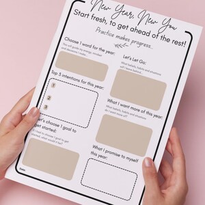 May include: A white paper with the text "New Year, New You" and prompts for goal setting. The prompts include "Choose 1 word for the year," "Top 3 intentions for this year," and "Let's Let Go." The paper is held by hands against a pink background.
