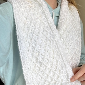Pode incluir: Uma echarpe branca, tricotada à mão, com um padrão de diamante texturizado. A echarpe é feita de um fio macio e espesso e tem uma borda ondulada. A echarpe é drapeada ao redor do pescoço e dos ombros.