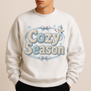 Op de afbeelding: Witte sweatshirt met de tekst "Cozy Season" in een lichtblauw en crèmekleurig ontwerp. Het ontwerp bevat een sneeuwpop, sneeuwvlokken en strikken. De sweatshirt heeft een ronde hals.