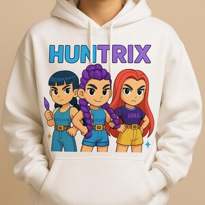 Op de afbeelding: Witte hoodie met het woord "HUNTRIX" in blauw en paars boven drie cartoonfiguren. De figuren hebben verschillende haarkleuren en outfits. Eén houdt een paars voorwerp vast. De hoodie heeft een capuchon met trekkoord en een kangoeroezak.