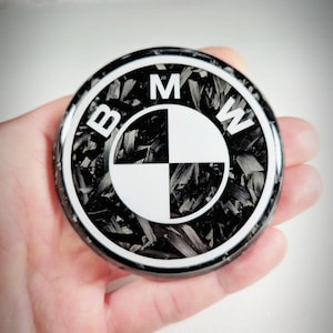 May include: A circular emblem featuring the BMW logo with a black and white checkered center and the letters BMW in white. The background of the emblem displays a textured, fragmented black and gray carbon fiber pattern with a glossy finish.