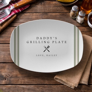 Puede incluir: Una fuente ovalada blanca con rayas verdes y blancas en los lados. El texto "DADDY'S GRILLING PLATE" y "LOVE, HAILEY" están impresos en negro. También se incluye un gráfico de tenedor y espátula cruzados.