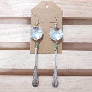May include: A pair of handmade silver spoon earrings. The earrings feature the bowl of a spoon as the top, with a green bead accent, and the handle of the spoon as the dangle. The earrings are displayed on a brown tag.