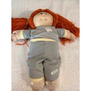 May include: A vintage Cabbage Patch Kid doll with orange yarn hair and blue eyes. The doll is wearing a gray long-sleeved shirt with a white cat graphic and gray pants. The doll's socks are beige and crocheted.
