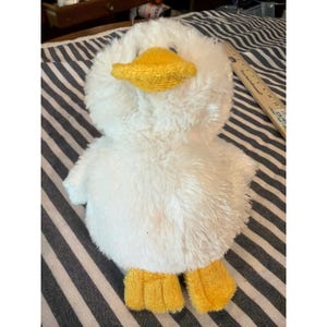 Op de afbeelding: Een pluche wit eendenspeeltje met een zachte, pluizige textuur, voorzien van een felgele snavel en bijpassende gele zwemvliezen. Het speelgoed ligt naast een houten liniaal met centimetervermeldingen voor de schaal en afmetingen.