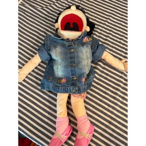 May include: A puppet with black hair, a wide open red mouth, and light skin tone. It wears a blue denim jacket with floral embroidery, light-colored pants, pink socks, and pink shoes with straps. The puppet has a button-down front on the jacket.