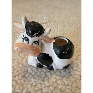 Może przedstawiać: Ceramiczna figurka krowy z błyszczącym białym wykończeniem, czarnymi łatami i jasnoróżowym pyskiem. Krowa ma duże oczy z długimi rzęsami, małe rogi i okrągły otwór na grzbiecie, zaprojektowany jako mała doniczka lub ozdobny pojemnik.