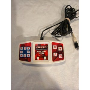 May include: A Coleco Handheld Video Game System controller. The white controller has red accents and black buttons. The text "COLECO Hand @ Hand VIDEO GAME SYSTEM" is printed on the front. A black cord and connector are attached.