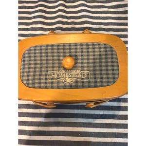 May include: An oval wooden basket with a light brown finish and a hinged lid. The lid features a blue and white checkered fabric insert with a central wooden knob and the embroidered text Longaberger Homestead in white thread.