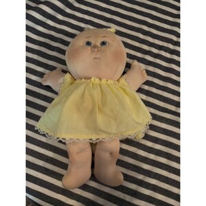 Könnte beinhalten: Eine Stoffpuppe mit rundem Gesicht, blauen Augen und rosigen Wangen. Die Puppe hat eine garnartige Haarstruktur auf dem Kopf und trägt ein hellgelbes ärmelloses Kleid mit einem zarten weißen Spitzenbesatz am unteren Saum des Kleidungsstücks.