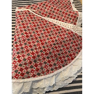 May include: A circular fabric tree skirt featuring a repeating grid pattern of red, green, and white squares with various holiday-themed illustrations. The edge is finished with a wide, white eyelet lace ruffle trim with a scalloped border.