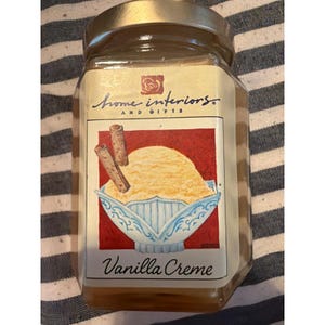 Pode incluir: Um frasco de vidro com tampa dourada contendo uma vela. O rótulo apresenta o texto Home Interiors and Gifts, uma ilustração de uma taça de gelado de baunilha com bolachas e o texto Vanilla Creme na parte inferior do rótulo.