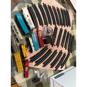 May include: A selection of model train parts, featuring colourful train carriages and black curved and straight tracks. The set includes a power supply and other accessories. The components are arranged on a pink surface, suitable for a model railway.