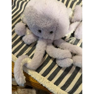 May include: A plush stuffed octopus toy made of soft, light purple faux fur. The creature features a rounded head, dark embroidered eyes, and eight long, flexible tentacles. A small white fabric tag with the text Bear is visible on one side.