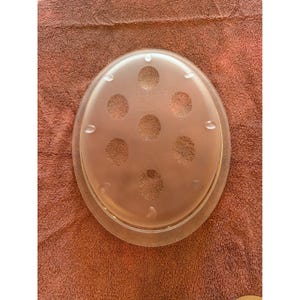 May include: A clear, oval-shaped Tupperware lid with six circular perforations and small raised bumps around the edge. The lid is made of a translucent plastic material and sits on a reddish-brown surface.