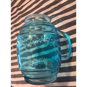 Op de afbeelding: Een tonvormige, lichtblauwe glazen pot met een handvat en een wijde opening. Het glas is aan de voorzijde voorzien van de in reliëf aangebrachte tekst FAMILY RECIPE in hoofdletters. De pot heeft een geribbeld, afgerond ontwerp met een gladde afwerking.