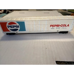 May include: A white model train car with the Pepsi logo in red, white, and blue on the side. The words "PEPSI-COLA" are printed in red on the side of the car. The car has black wheels and is on a light gray surface.