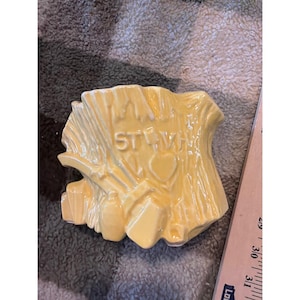 Può includere: Un pezzo decorativo in ceramica gialla con le parole "I'M ST. WH" e un motivo a cuore. Il pezzo ha un aspetto strutturato simile al legno e un piccolo elemento a forma di bottiglia sul lato.