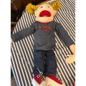 May include: A puppet with yellow yarn hair in pigtails, a beige face, and a wide red mouth. It wears a long-sleeved navy and white striped shirt with the red text "SUNNY GIRL" embroidered on the chest, dark blue denim pants, and red sneakers.