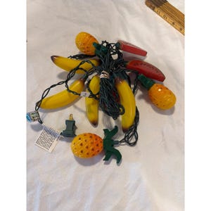 May include: A string of novelty lights featuring yellow bananas, orange pineapples, and red watermelon shapes. The lights are strung on a green wire with a standard plug. The lights are designed to look like tropical fruits.