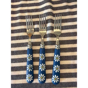 Può includere: Tre forchette d'argento con manici blu decorati con motivi a margherita bianchi e gialli. Le forchette sono disposte su un tessuto a righe, mettendo in risalto il design decorativo delle posate.