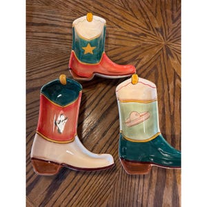 May include: Three ceramic dishes shaped like cowboy boots. One boot features a yellow star on a teal background. Another displays a small white horse head graphic on a red and teal surface. The third boot shows a tan sombrero illustration.