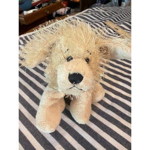 Puede incluir: Un perro de peluche con pelaje beige claro desgreñado, orejas caídas y ojos de plástico negro. El juguete tiene una nariz y boca cosidas en negro, con una textura suave en el cuerpo y las patas, midiendo aproximadamente 15 cm de altura.