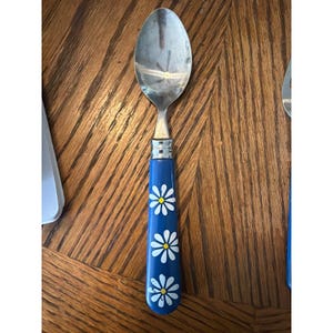 May include: A stainless steel spoon featuring a blue handle decorated with three white daisy flowers with yellow centers. The handle has a smooth, cylindrical shape and is connected to the metal bowl by a silver-colored metal ferrule.
