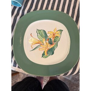 May include: A square ceramic plate with a wide, solid sage green border and a cream-coloured centre. The centre features a hand-painted illustration of yellow lily-like flowers with green leaves, rendered in a painterly style with visible brushstrokes.