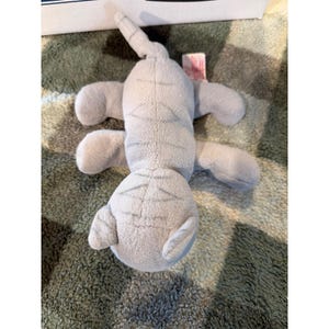 May include: A light grey plush cat toy featuring a soft, fuzzy texture and darker grey stitched stripes along its back, head, and tail. The stuffed animal is shown from a top-down perspective, displaying four legs, a small tail, and ears.
