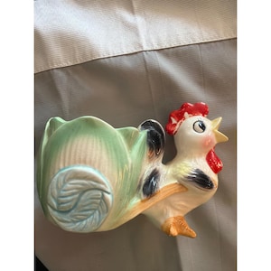 May include: A ceramic planter in the shape of a rooster. The planter has a light green, leaf-shaped base and a white rooster with red comb and wattle. The rooster is perched on a branch and has black and yellow accents.