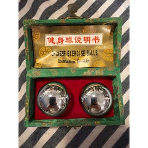 Peut inclure: Une paire de boules d'exercice en chrome poli logées dans une boîte recouverte de tissu vert avec une doublure intérieure rouge. Le couvercle présente un insert doré avec le texte "CHINESE EXERCISE BALLS Instruction Booklet" et des caractères chinois.