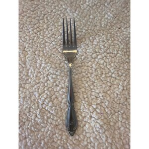 May include: Silverware fork with four tines and a decorative handle. The handle has an engraved design and a monogrammed initial. The fork is made of a shiny, reflective metal.