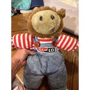 May include: A plush stuffed animal with brown fur, wearing a red and white striped shirt under blue denim-style overalls. The overalls feature two red buttons at the straps and a rectangular patch on the chest with the white text BI LO.