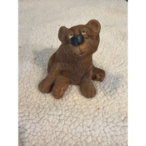 May include: A brown bear figurine with a smiling expression and blue eyes. The bear is sitting with its paws outstretched. The nose is black. The figurine is made of a textured material.