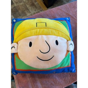 May include: A plush square pillow featuring a cartoon face with a yellow hard hat, a peach-colored face, and a smiling expression. The pillow has a blue border and is designed for children.