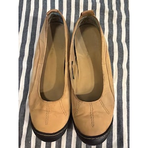 May include: A pair of tan slip-on shoes featuring a rounded toe, visible tonal stitching details on the upper, and a dark brown rubber sole. The interior footbed is tan with a visible brand logo, and the overall design is a low-profile flat shoe.