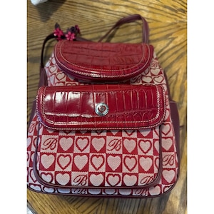 Peut inclure: Un sac à dos rouge et beige avec un motif répétitif de cœurs et de la lettre B. Le sac possède un rabat et une poche avant en similicuir rouge brillant façon croco, ornés d'un emblème circulaire argenté avec un cœur et la lettre B.
