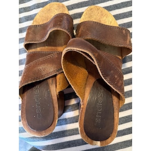 May include: Pair of brown leather Candie's clogs with wooden soles. The clogs have a strap across the top of the foot and a wooden base. The footbed is a darker brown color. The clogs are on a striped background.