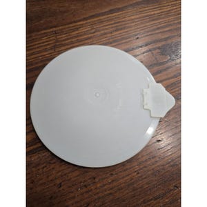 May include: A round, semi-translucent white plastic Tupperware lid featuring a centred circular indentation and a small, protruding tab on the right side. The surface is smooth with subtle embossed text visible near the centre point.