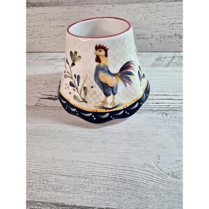 May include: A ceramic rooster-themed candle holder. The holder features a hand-painted rooster with blue, yellow, and red feathers. The base has a navy blue border with white dots. The interior rim is red.