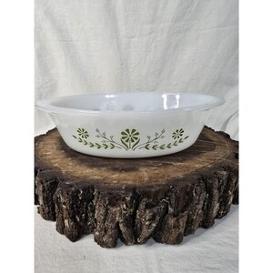 May include: White oval serving bowl with a green floral pattern. The bowl is made of glass and sits on a rustic wooden surface. The floral design includes stylized flowers and leaves, adding a vintage touch to the kitchenware.