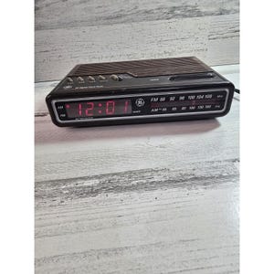 Puede incluir: Radio reloj digital GE negro con una pantalla LED roja que muestra las 12:01 AM. La radio tiene opciones de frecuencia de radio FM y AM. La parte superior tiene una rejilla de altavoz marrón y botones.