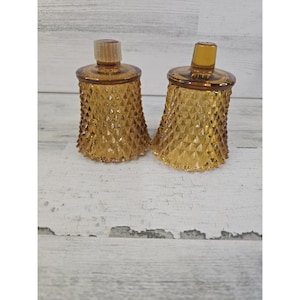 May include: Two amber-colored glass votive candle holders featuring a raised diamond pattern across the exterior surface. Each piece has a smooth, circular rim at the top and a small, cylindrical neck extending upward from the center.