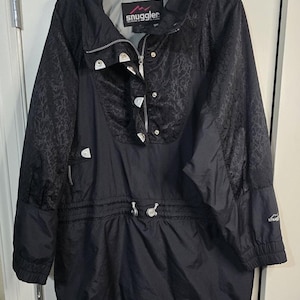 May include: Black Snuggler anorak jacket with a high collar, zipper closure, and decorative floral pattern on the upper portion. Features a drawstring waist and the Snuggler logo on the sleeve. A practical and stylish outerwear garment.