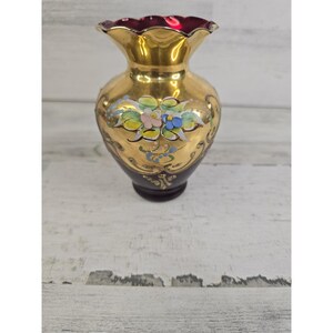 Puede incluir: Un pequeño jarrón de cristal con una base de color rojo arándano intenso y una sección superior dorada. El jarrón presenta un borde festoneado y está decorado con motivos florales pintados a mano en tonos rosa, azul, verde y blanco, junto a volutas doradas.